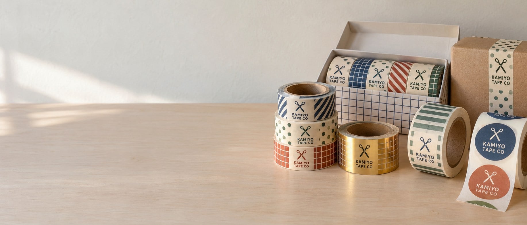 Custom printed, foil and wide washi tape rolls stacked beside a gift set and a taped kraft box against a white wall
